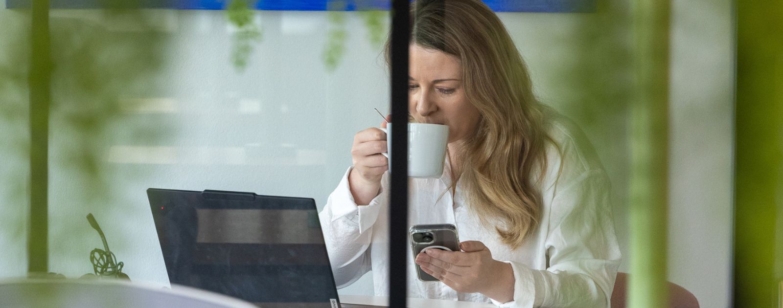 A woman sipping from a cup while working on her laptop and smart phone.