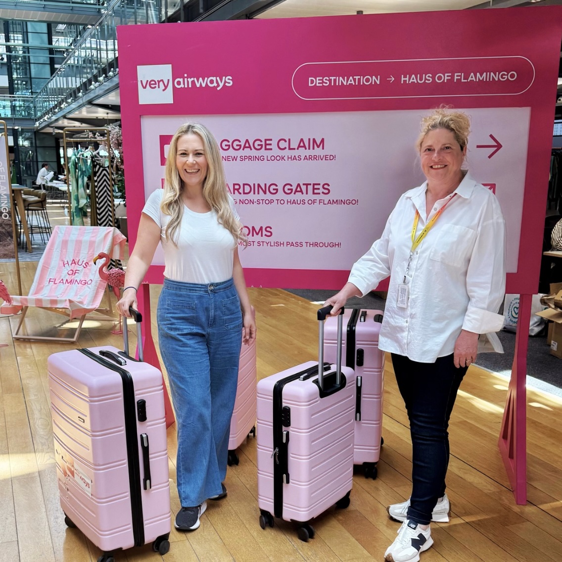 Two colleagues holding luggage bags and standing in front of a Very Airways display.